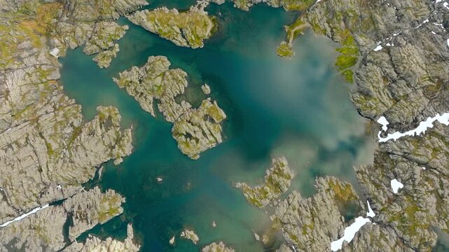 Aerial drone footage looking straight down over a small high-altitude mountain lake surrounded by rugged rocky terrain and patches of snow along the Trolltunga hiking route in Norway. The drone moves 