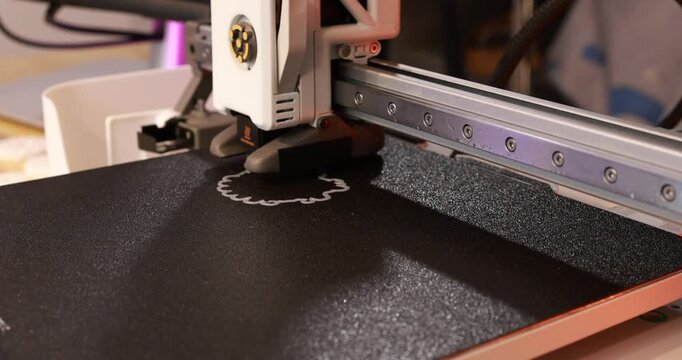 Close-up of a 3d printer extruding material on a black build bed, the print head and rail in a workshop setting.