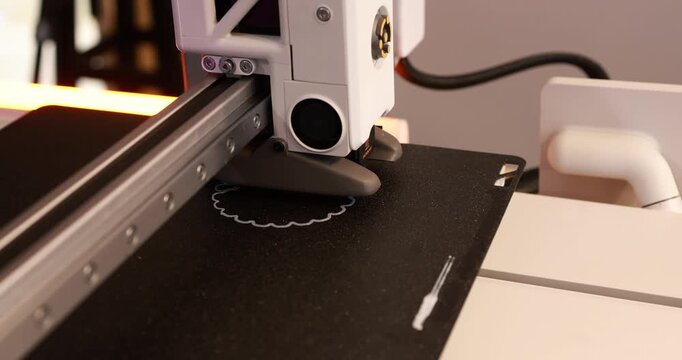 Close-up of a 3d printer head printing a small object on a black build plate in a workshop.