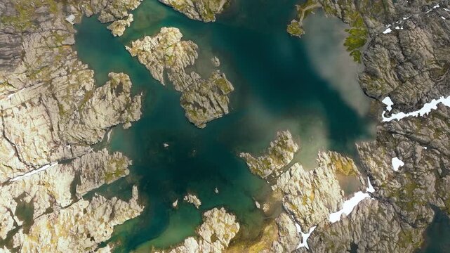 Aerial drone footage looking straight down over a small high-altitude mountain lake surrounded by rugged rocky terrain and patches of snow along the Trolltunga hiking route in Norway. The drone moves 