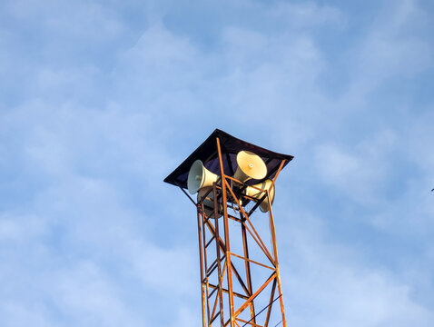 Loudspeaker horn tower mounted high against a cloudy blue sky.