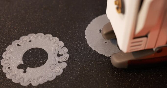 Close-up of a 3d printer creating a gray decorative ornament on a dark surface, with a finished printed piece nearby.