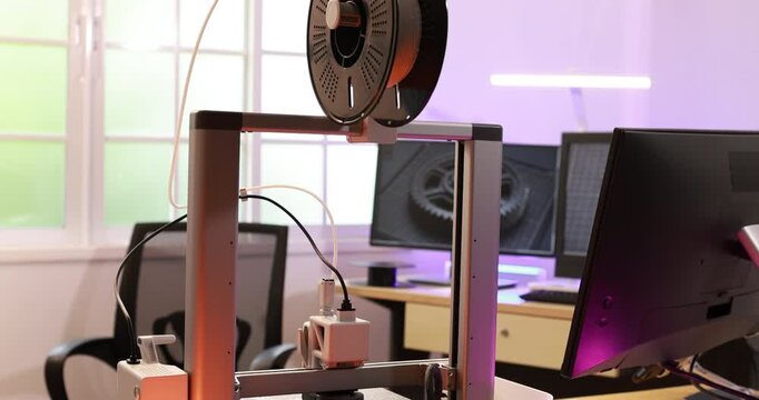 3d printer with filament spool in a modern office workspace with computer monitors and soft purple lighting.