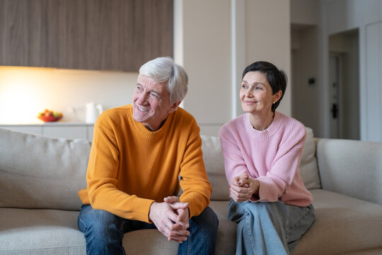 Happy senior married couple husband and wife sitting couch at home looking away at window, smiling elderly man and woman talking having fun together and spending time in good mood, family life harmony
