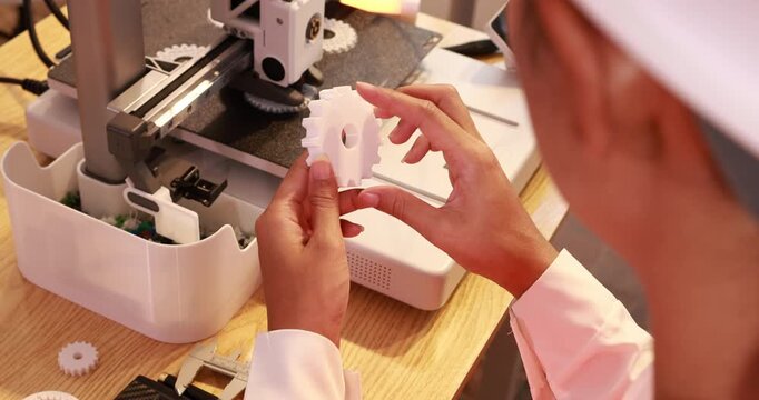 Hands holding a 3d printed gear beside a desktop 3d printer in a workshop, prototype making and engineering work.