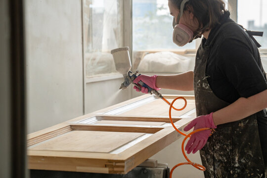 Craftswoman painter wearing protective respirator mask paints antique wooden door with spray gun, female master in safety equipment using instrument in paint booth, spraying paint evenly over surface.