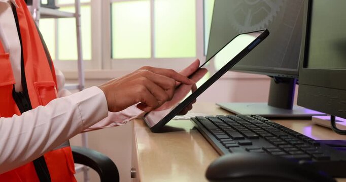 Person in safety vest using a tablet at a desk with computer and keyboard in a bright office setting.