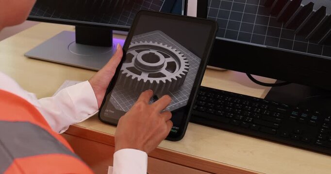 Person using a tablet to inspect a gear design at a desk with computer monitor and keyboard in an office or workshop