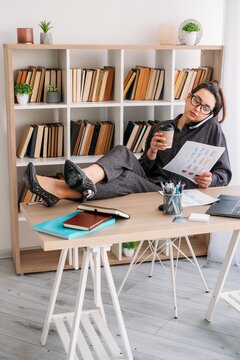 Business call. Office routine. Female manager talking on smartphone drinking coffee at desk with notebook analyzing paperwork at professional workplace.