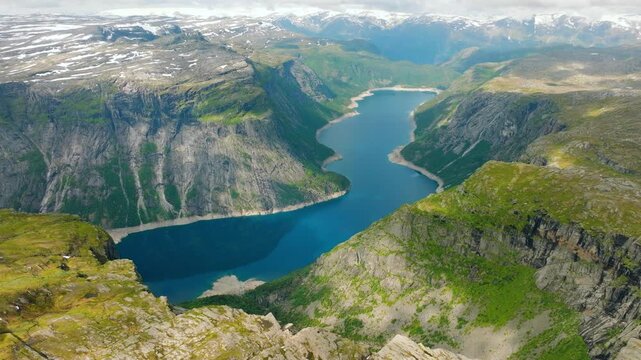 Cinematic aerial drone footage of Ringedalsvatnet, a dramatic mountain reservoir surrounded by steep rocky cliffs and lush green slopes in western Norway. The famous Trolltunga overlooks the reservoir