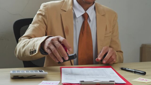 Professional in suit stamps tax document at office desk with calculator and notes