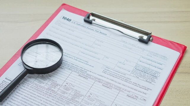 Wooden tiles spelling tax sit next to magnifying glass on top of 1040 tax form clipboard