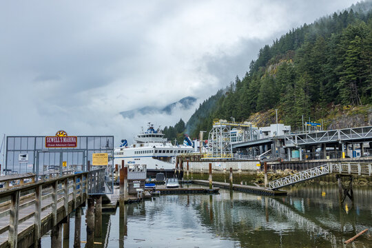 A BC Ferries vessel berths at the terminal beside a gated Sewell's Marina boardwalk, a metal gangway dropping to floats as cloud spills over the forested ridge.