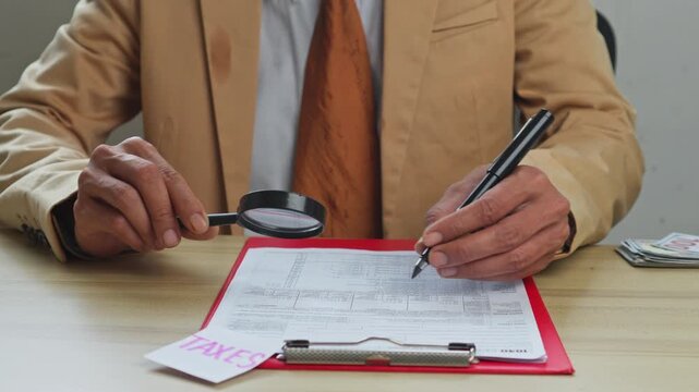 Professional in suit uses magnifying glass and pen to review and fill out tax forms on desk