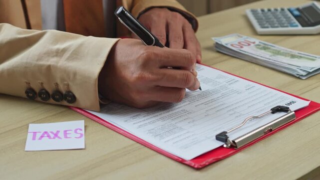 Person in suit fills out tax return forms with pen next to money and calculator