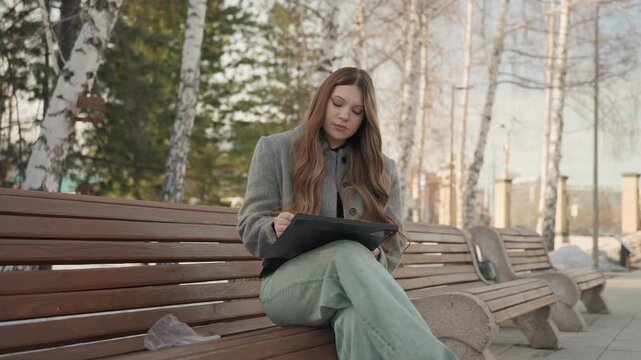 Elegant woman writing. Woman engrossed in script outdoors. Thoughtful lady reviewing notes on park bench. Poised woman surrounded by nature while contemplating her screenplay notes in winter