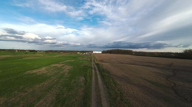 Wide aerial view tracking forward over a narrow dirt road separating a green grassy pasture and a raw cultivated farm field towards a coverall under a dramatic cloudy blue sky in Milton Ontario.