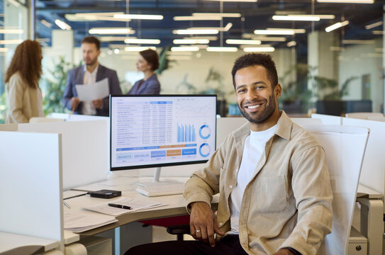 Analyst man in office with computer dashboard. Smiling professional at desk reviews charts and report as colleagues talk nearby, underscoring business finance teamwork. Key idea is team data.