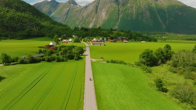 Aerial drone flight along Route 63 through a lush Norwegian mountain valley. The camera moves forward along the road while gradually ascending, revealing green fields, forests, rural landscape 