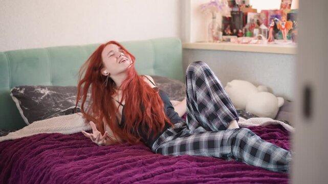 Brunette woman relaxing. Redheaded Caucasian woman stretching on colorful bedding in morning light. Freckled redhaired woman relaxes on purple bed surrounded by cozy blankets and childhood dolls