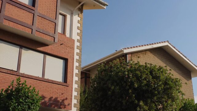 Modern residential buildings with brick facades and tiled roofs under a clear blue sky, featuring green trees and warm sunlight