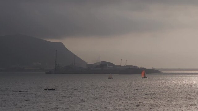 Sailboats crossing tranquil bay waters under a cloudy sky with industrial port structures and distant mountains creating an atmospheric scene