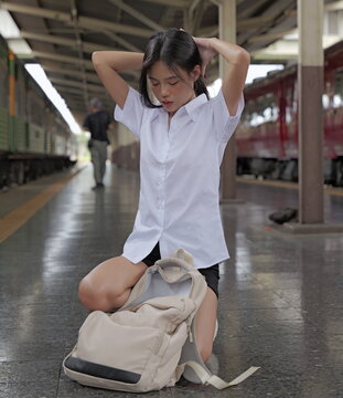 A young Asian woman in a university student uniform kneels on a train station platform next to an open backpack, adjusting her hair with both hands raised behind her head