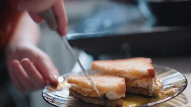 White woman slicing runny egg sandwich, knife breaking golden yolk that oozes across plate, cinematic closeup capturing motion, glistening texture, candid home kitchen indulgence and playful messy