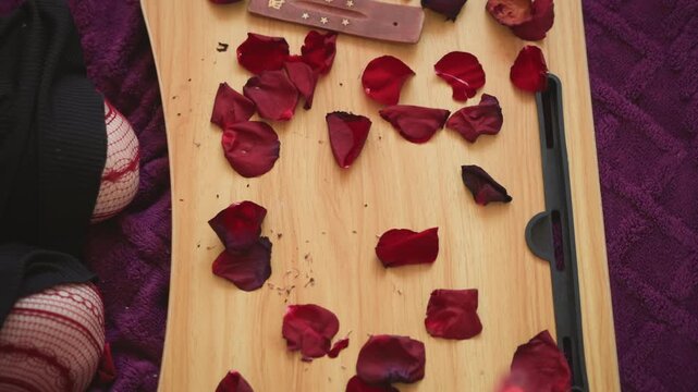 Rose petals scattered on tray, mystic reader arranges petals with wooden wand over wooden board, tarot cards visible at edge, purple cloth backdrop, closeup hands, sensual atmospheric ritual for love