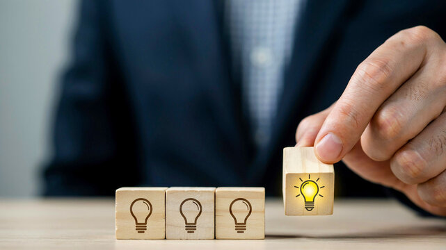 Person placing a wooden block with a lit lightbulb icon onto a line of blocks with unlit lightbulb icons, symbolizing innovation and new ideas