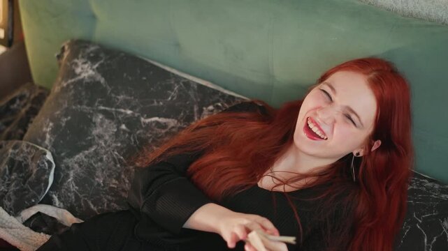 Red haired caucasian woman laughing on sofa holding tarot cards, cozy green couch with marble cushion, bohemian jewelry and necklace, warm ambient light, relaxed fortune telling session, playful