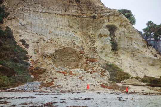 A steep coastal bluff shows evidence of a recent landslide with orange safety cones placed on the sand, North Pacific beach, San Diego, CA
