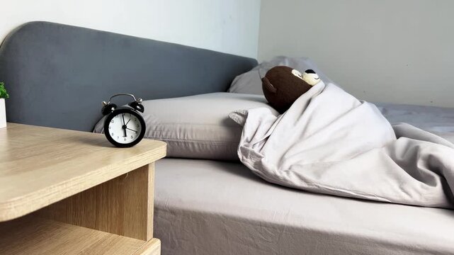 In the bright bedroom, a teddy bear sleeps under a gray blanket on a soft pillow, a black alarm clock ticks on a wooden bedside table, and a plant turns green. The concept conveys the comfort of home