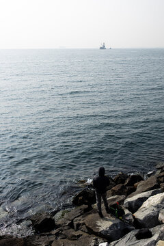 Fisherman stands on rocks by the sea while a boat sails in the distance