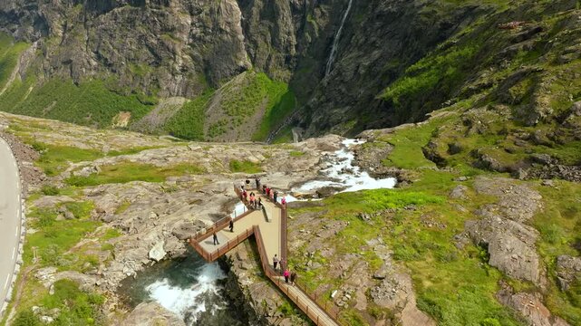 Smooth cinematic drone footage of a scenic viewpoint near Trollstigen in Norway, showing the elevated pedestrian walkway winding above a mountain stream and rocky alpine terrain. Tourists explore 