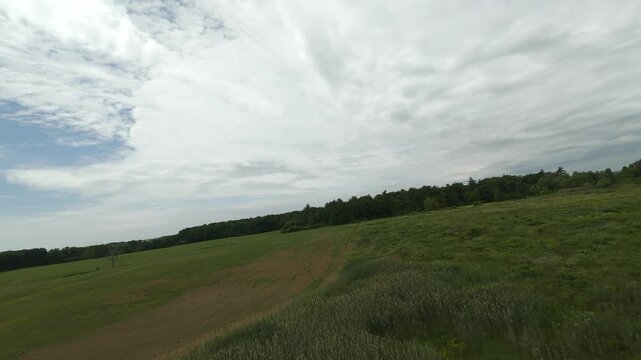 Panoramic drone shot pans across rolling green agricultural fields bordered by thick summer woods under a wide blue sky filled with white clouds.
