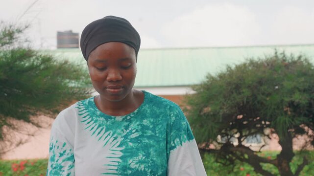 Black woman outdoors contemplative gaze, caregiver portrait with teal patterned shirt and headscarf, soft sunlight and green bushes with pale roof background, gentle downward glance and serene mood,