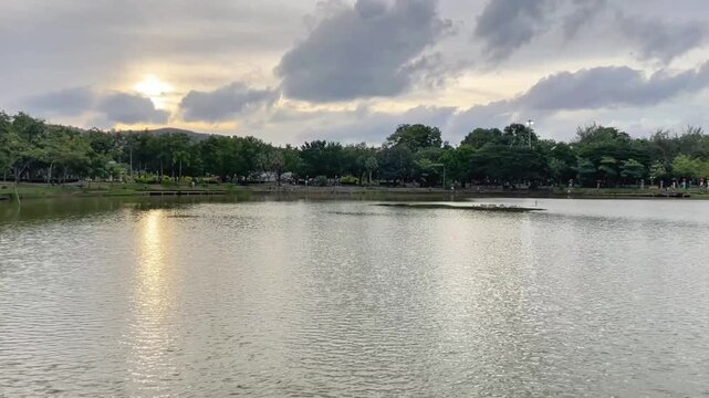 A tranquil lake at sunset in King Rama IX Park in Phuket, Thailand. Sunlight reflects off the water, while the cloudy sky and green vegetation create an atmospheric tropical landscape.