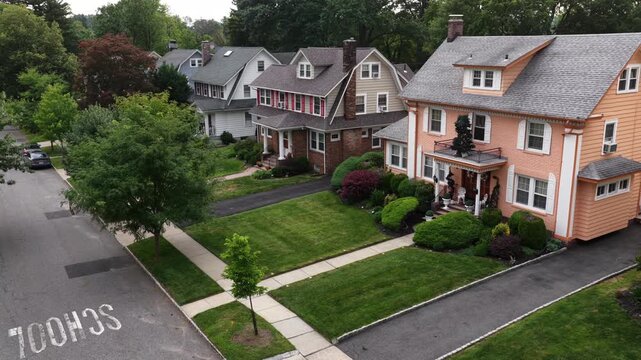 Peach colored house with balcony porch stands beside neighboring homes on tree lined street in South Orange, New Jersey, sidewalks and manicured lawns border quiet suburb. Aerial drone view.