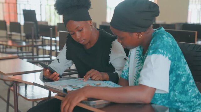 Classroom black mother and child studying together at wooden desk with workbook and smartphone, mother tutoring daughter, warm supportive interaction, focused concentration, soft sunlight