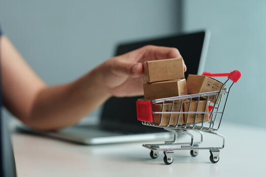Woman placing small packages into a miniature cart beside a laptop, representing e-commerce orders, online shopping, product delivery, digital retail, logistics, and fulfillment.