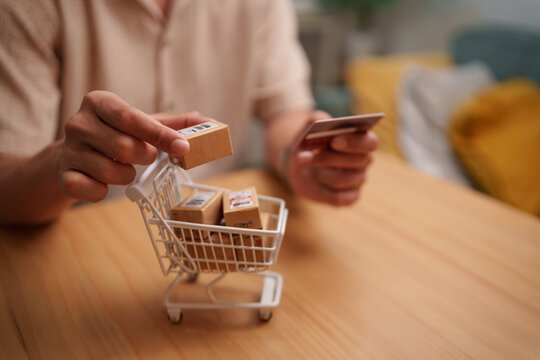 Asian Man holding a payment card and parcel above a miniature shopping cart, representing e-commerce, online payment, delivery, digital retail, purchasing, and consumer convenience.