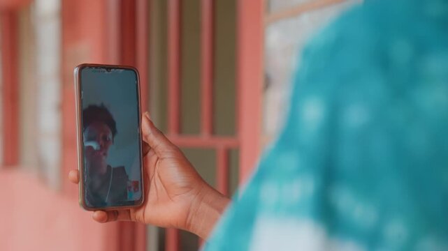Black mother viewing child on phone, closeup of hand holding smartphone with childs reflection on screen turquoise sleeve, doorway backdrop, tender laugh and surprised gesture, heartfelt remote