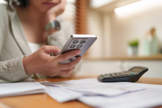 Asian Woman checks financial information on a smartphone beside bills and a calculator, managing expenses, budgeting, and personal finances for smarter everyday money decisions.