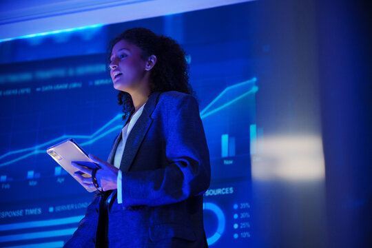 Professional woman giving a business presentation with a tablet beside a digital network analytics dashboard, showing traffic data, charts, performance metrics, and technology insights.