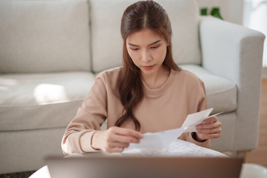 Concerned Asian woman reviewing household bills and receipts at home, calculating monthly expenses and managing personal finances, budget, payments, savings, and financial planning.