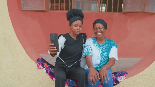 Black mother and daughter sitting bench taking selfie smartphone in urban courtyard, colorful textile cushion, playful smiles, family bonding, mother caregiver capturing memory while daughter giggles
