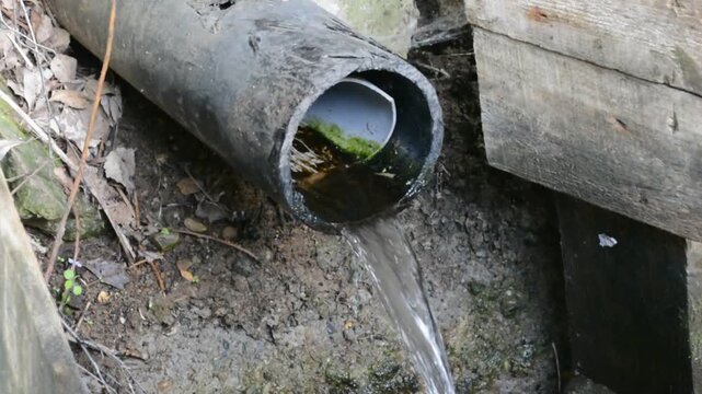Water flowing from a black plastic pipe in a dirt landscape. Stream of liquid draining from conduit into ground. Drainage system maintenance or sewage runoff.