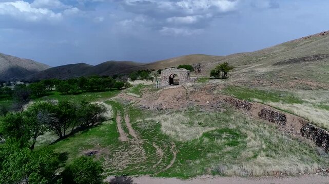 Aerial drone footage captures ancient stone fire temple ruins standing on green hillside with surrounding mountains and valley under bright daylight showcasing historical zoroastrian heritage site.