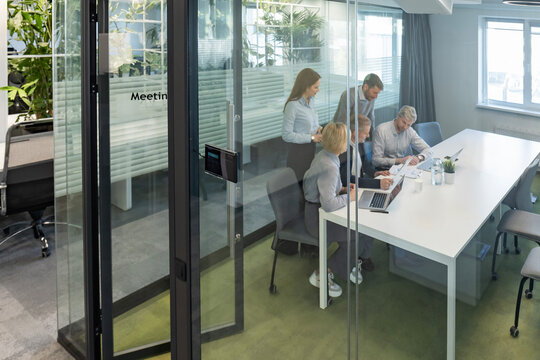 A view through a glass partition shows a modern, busy office interior. Business people are working at desk